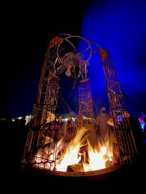 A large fire sculpture with flames rising from a metal fire pit, surrounded by people at night, from Fire & Story 2026 on the Waypoint Park and Trackside grounds.